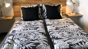 Una cama con cabecera de madera, dos almohadas con estampado blanco y negro, y una mesita de noche con una lámpara. Encima de la cama, hay tres cuadros enmarcados en la pared.