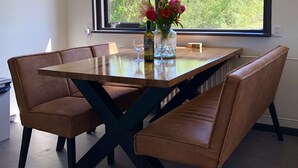 Un comedor con una mesa y bancos, un jarrón con flores, y una ventana con vista a árboles.