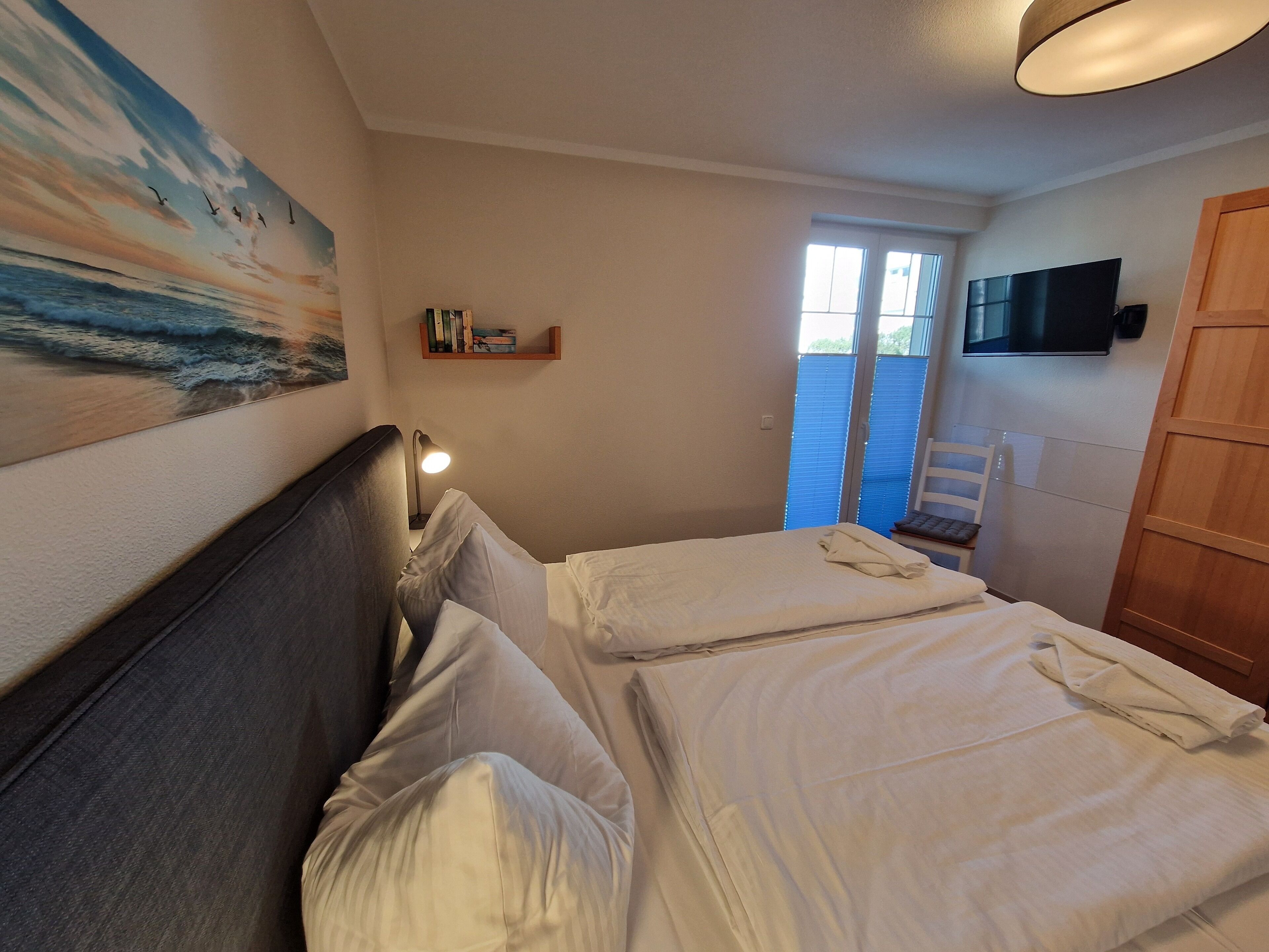 Ein Schlafzimmer mit zwei Betten, einem Nachttisch mit Lampe, einem Bücherregal, einem Fernseher und einem Fenster mit Jalousien.