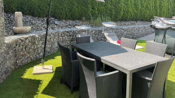 Een terras buiten met een tafel en stoelen, een stenen fontein en een haag op de achtergrond.