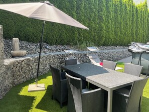 Un patio esterno con tavolo e sedie, una fontana in pietra e un siepe sullo sfondo.