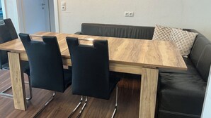 Een eetruimte met een houten tafel en zwarte stoelen, een bank met een gekleurd kussen, en een lichtgekleurde muur met stopcontacten.