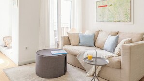 A modern living room with a beige sofa, a round ottoman, a small side table with a glass of orange juice, a map on the wall, and a large window with white curtains. - Modern comfort in Föhr's prime location (Nieblum)