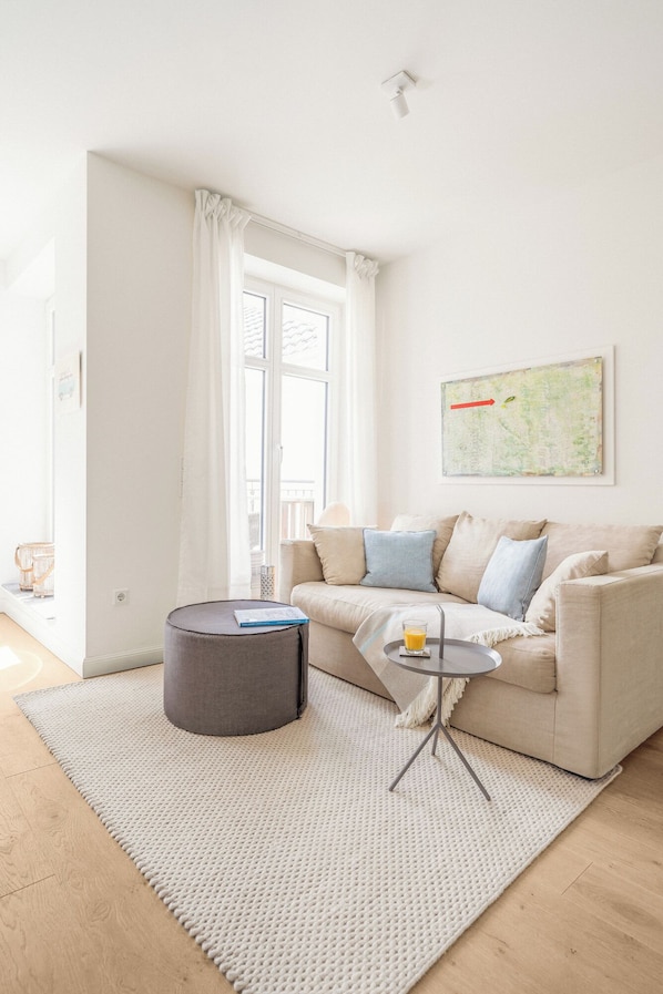 A modern living room with a beige sofa, a round ottoman, a small side table with a glass of orange juice, a map on the wall, and a large window with white curtains. - Modern comfort in Föhr's best location (Nieblum)