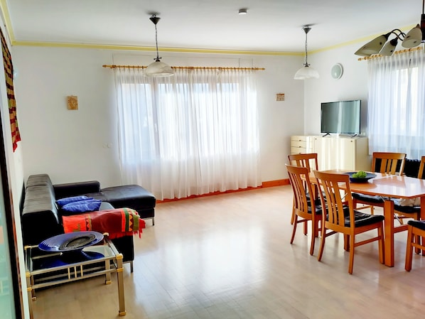 Una sala de estar con un sofá, una mesita con un ventilador, un comedor con una mesa y sillas, y un televisor sobre un mueble.