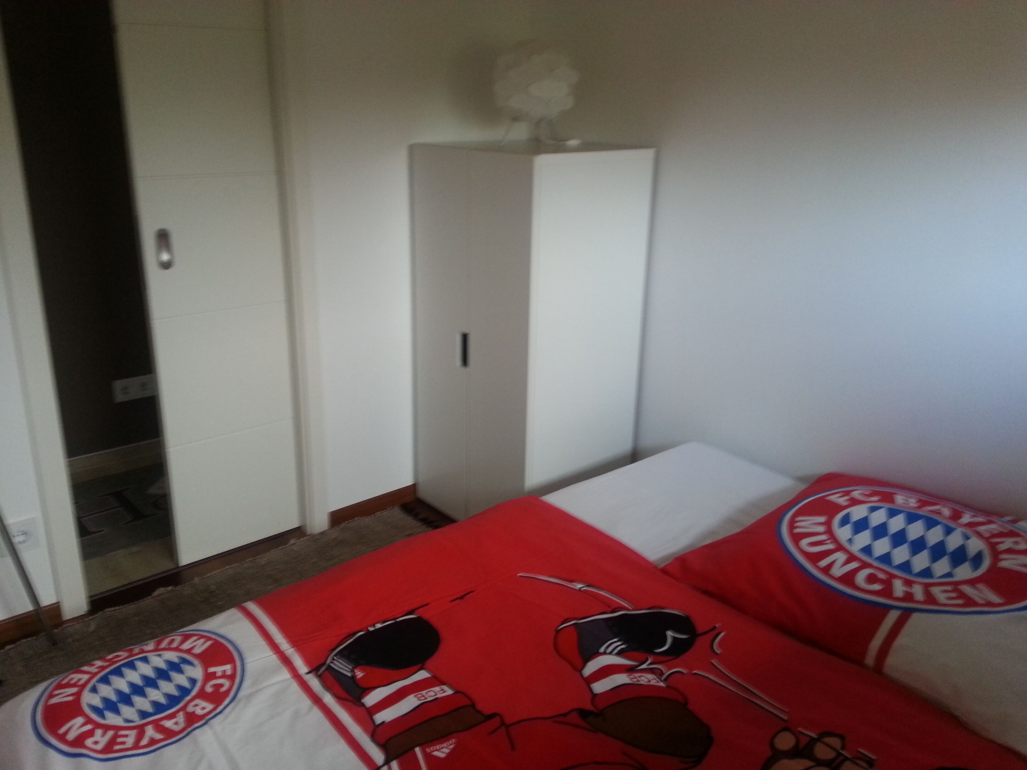 Een slaapkamer met een bed, voorzien van kussens van FC Bayern München, een witte inbouwkast en een deur naar een andere kamer.
