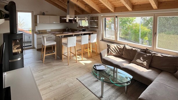A spacious loft with a high ceiling, wooden beams, a kitchen area with a dining table and chairs, a sofa, and large windows offering a view of the outdoors.