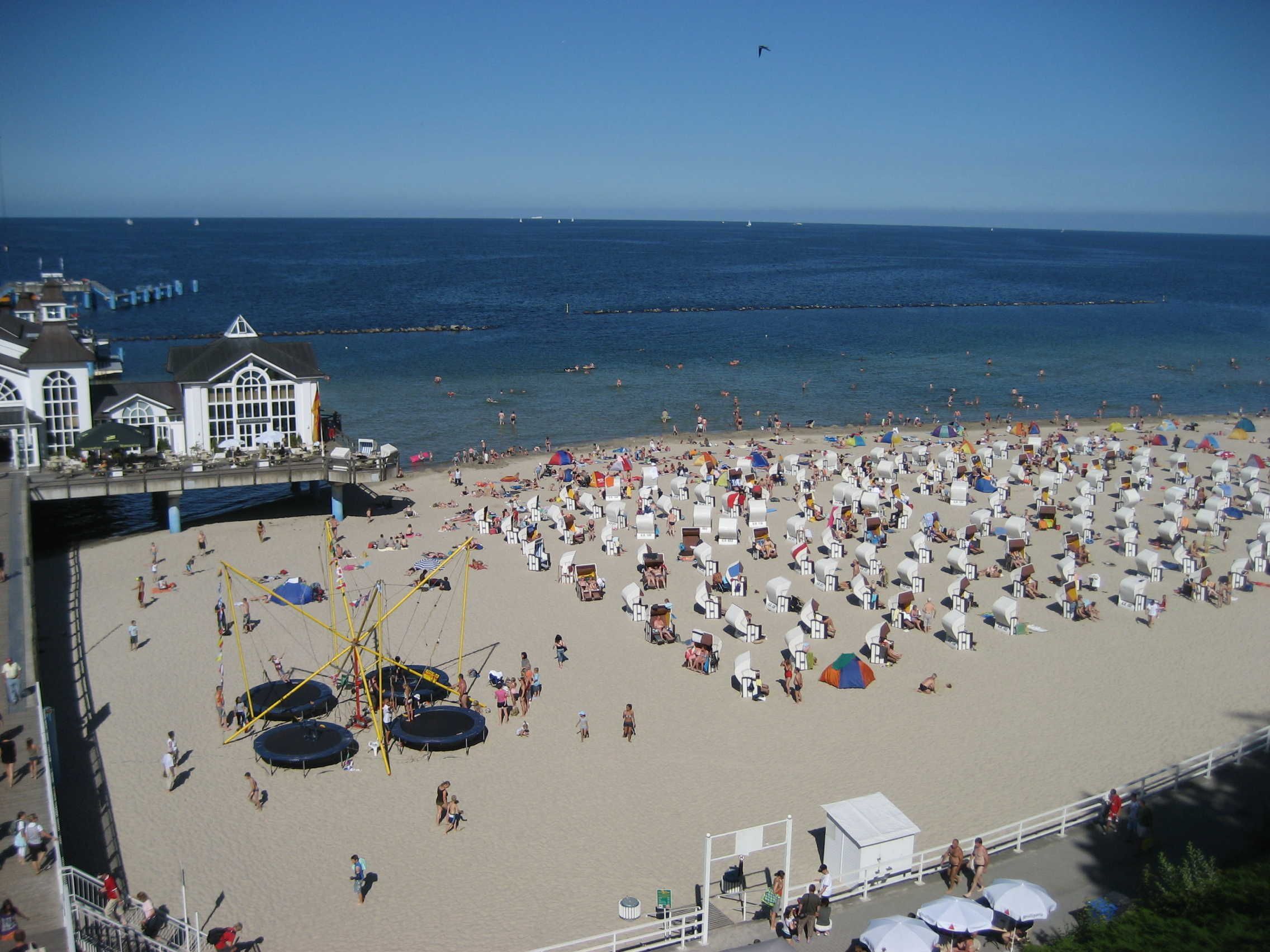 Une plage bordée de nombreux parasols et de chaises longues, une jetée avec des bâtiments, et l’océan en arrière-plan.
