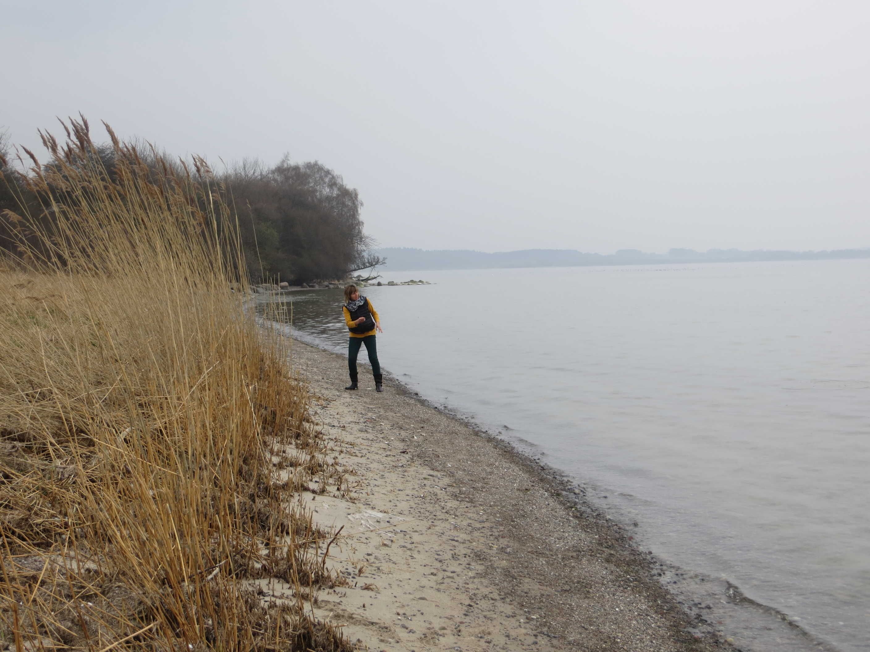 A person standing by a calm body of water with dry grass and trees in the background.