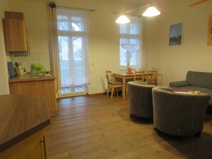 Una habitación con piso de madera, una mesa de comedor con sillas, un sofá, una zona de cocina con mostrador y armarios, y una ventana con cortinas.