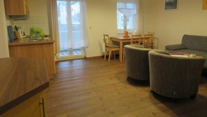 A room with a wooden floor, a dining table with chairs, a sofa, a kitchen area with a counter and cabinets, and a window with curtains. - Wittow - Pension Waldrose (Sellin)
