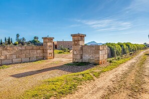 Eine Steinmauer mit einem Tor, ein Feldweg und ein Zaun mit Grünpflanzen.