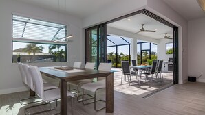 Un espace repas moderne avec une table en bois et des chaises blanches, de grandes fenêtres et une vue sur une piscine et un coin salon extérieur.