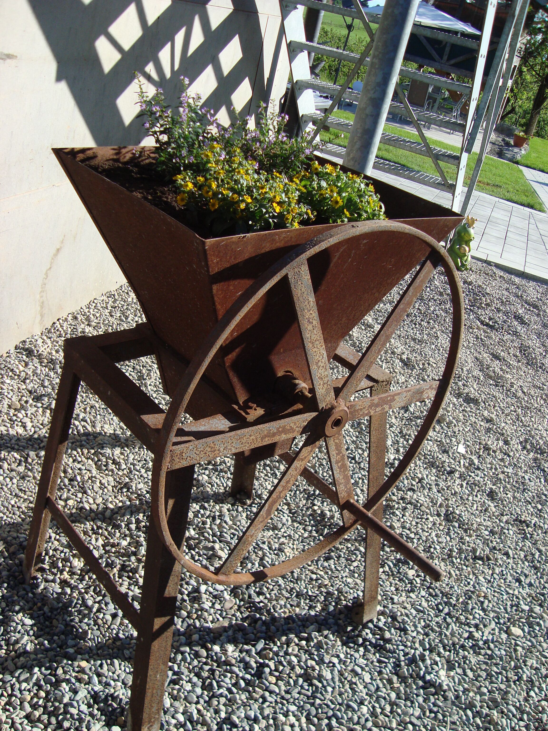 A rusted metal wheelbarrow with a flower planter on a gravel surface.
