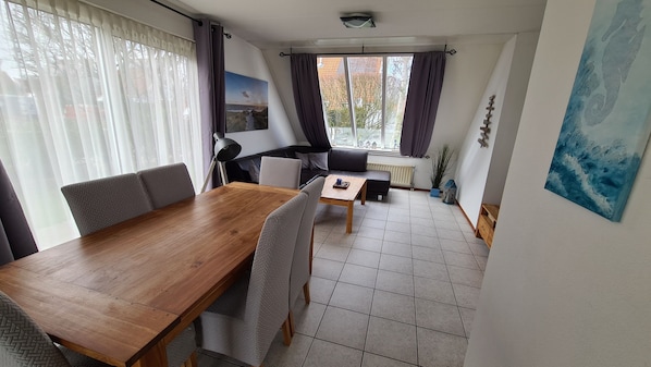 A modern living room with a wooden dining table, grey upholstered chairs, a black sofa, and a view of the outdoors through large windows.