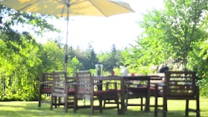 Un espace extérieur aménagé avec des tables et des chaises en bois, un grand parasol et une végétation luxuriante en arrière-plan.