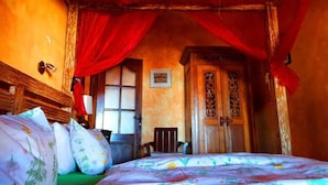 Una stanza con un letto, una testiera in legno, una tenda rossa, una porta in legno finemente intagliata e un tavolino con una sedia.