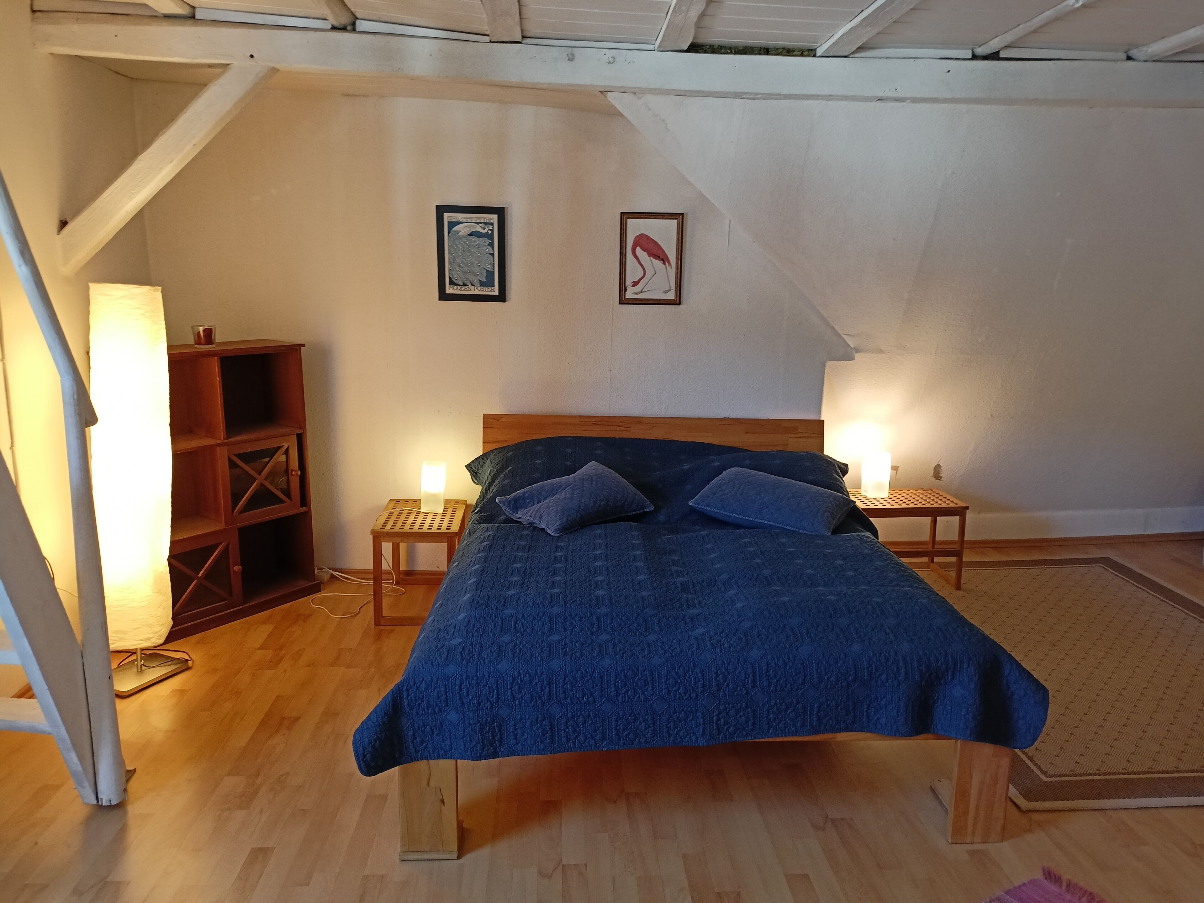 Ein Schlafzimmer mit einem Bett, zwei Nachttischen, einem Holznachtkästchen, einem Regal und zwei gerahmten Bildern an der Wand.