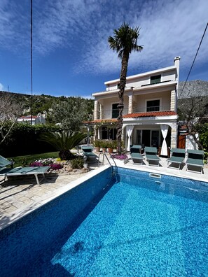 A two-story building with a swimming pool, outdoor seating, and a mountain backdrop. - Beachfront Villa Omiš – 20m from Sea, Heated Pool, Sauna & Top Location (Omis)