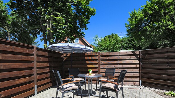 A patio with a table and chairs, a striped umbrella, and a wooden fence. - Non smoking apartment, ideal for 2 to 4 persons, quiet place suitable for allergy sufferers, (Stedesdorf)