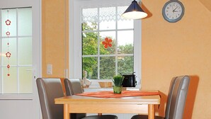 A dining area with a wooden table and four chairs, a hanging pendant light, a clock on the wall, and a window with a view of greenery. - Non smoking apartment, ideal for 2 to 4 persons, quiet place suitable for allergy sufferers, (Stedesdorf)