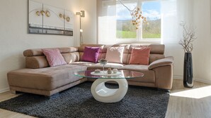 Un salon moderne avec un canapé d’angle, une table basse en verre et une grande fenêtre donnant sur un paysage verdoyant.