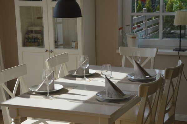 A dining table set with white chairs, a black pendant lamp, and a window with a view of greenery.