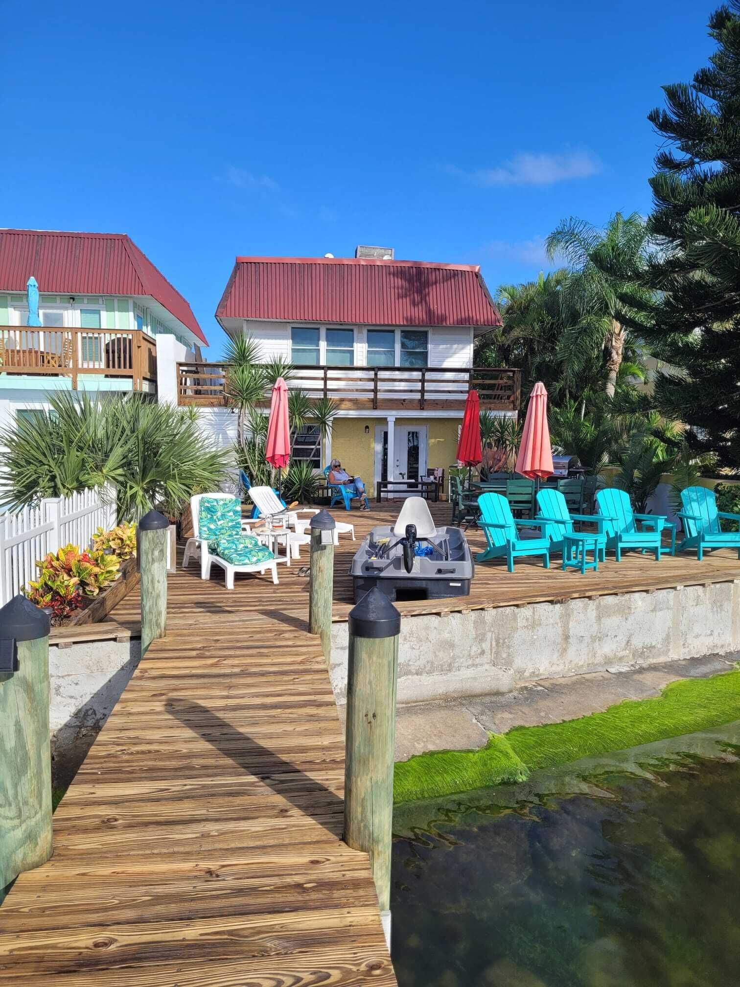 A wooden deck leading to a building with a red roof, outdoor seating with chairs and umbrellas, and a small boat.