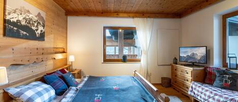 A cozy wooden cabin room with a bed, a dresser, a television, and a window with a view of a mountain.
