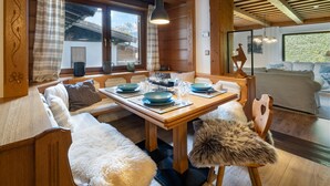Uma área de refeições acolhedora com uma mesa de madeira posta para quatro pessoas, bancos com mantas de pele e vista para uma cabana na montanha através de amplas janelas.