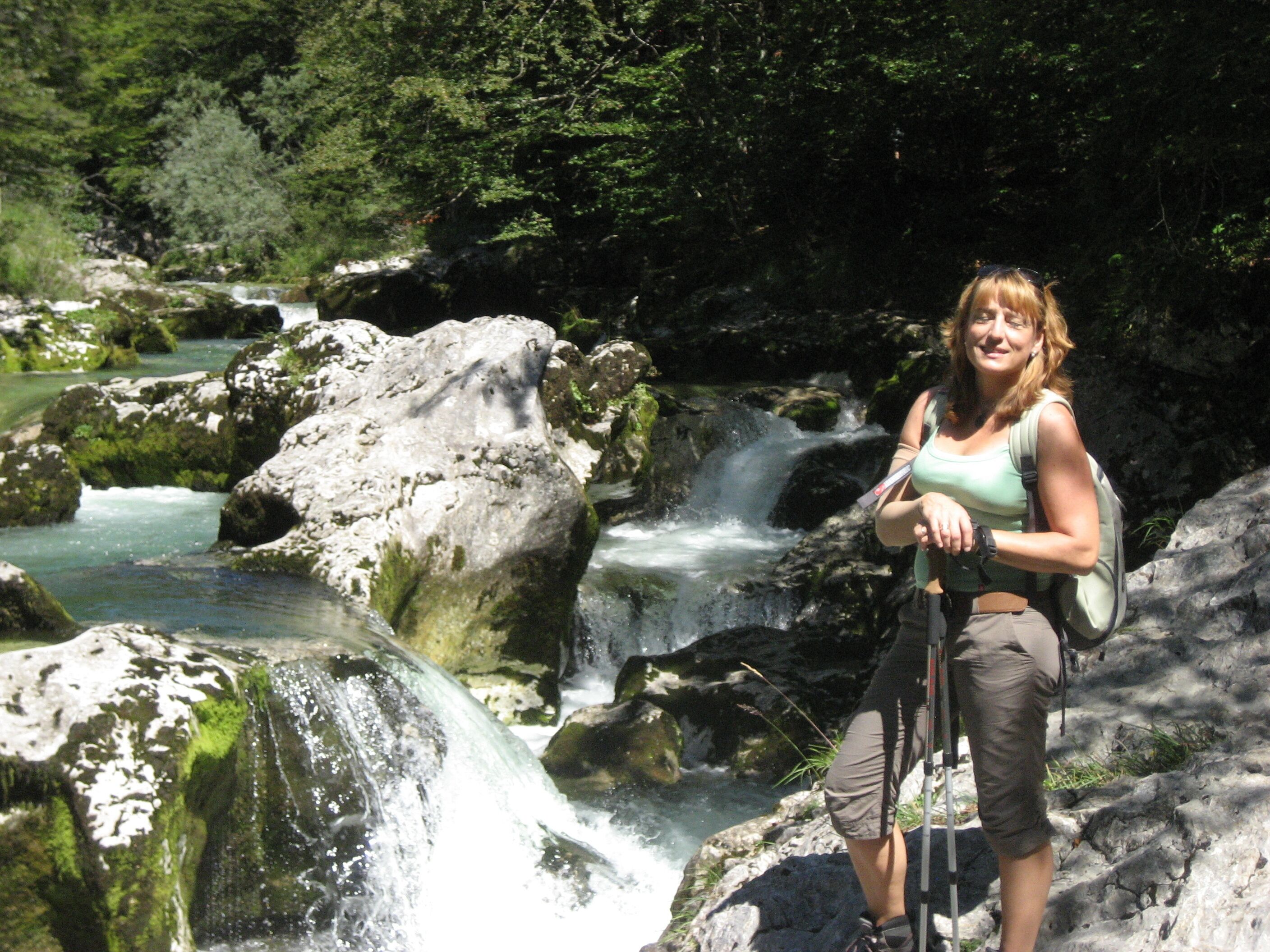 A woman standing by a waterfall with a backpack, surrounded by rocks and greenery.