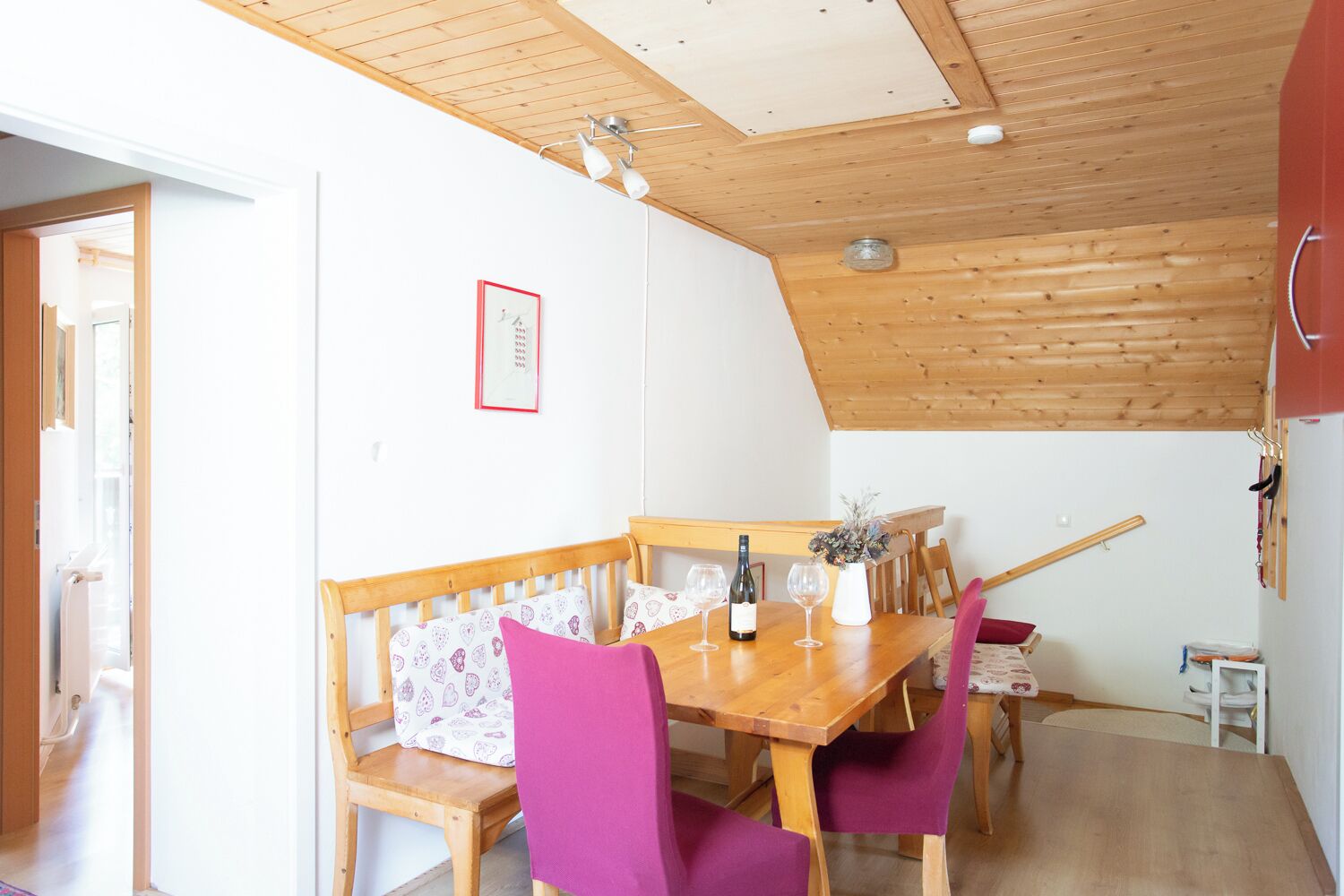 Un coin repas avec une table en bois, des chaises violettes et un banc. On peut voir une bouteille de vin et un vase rempli de fleurs sur la table.