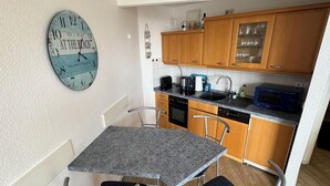 Une cuisine compacte avec des meubles en bois, un plan de travail en granit, un évier et un four à micro-ondes. Il y a une horloge au mur et une petite table à manger avec deux chaises.