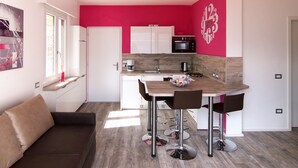 Een moderne keuken met witte kasten, een houten aanrecht en een roze accentmuur. Er is een zithoek met een tafel en stoelen, en een bruine bank in de hoek.