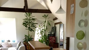 Un salon moderne avec une table basse en bois, un tabouret de bar et une grande plante en pot. On aperçoit un canapé et une table à manger à l’arrière-plan.