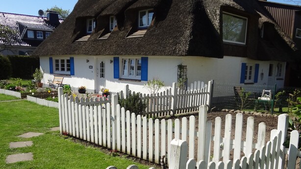 Une maison traditionnelle à toit de chaume, avec une clôture en bois peinte en blanc, une pelouse bien entretenue et un ciel bleu dégagé.