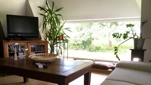 Un salon avec une table basse en bois, un téléviseur, une bibliothèque et une grande fenêtre donnant sur un paysage verdoyant.
