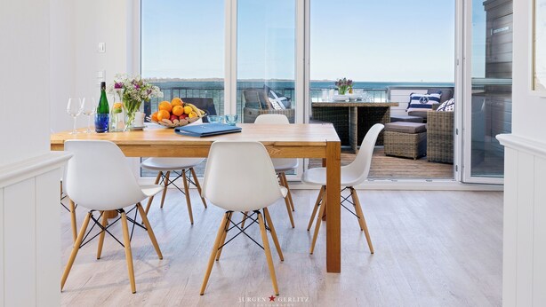 Un coin repas avec une table en bois, des chaises blanches et une vue sur la mer, visible à travers de grandes portes vitrées.