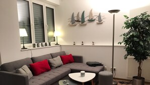A modern living room with a grey sectional sofa, red and grey cushions, a small round table with two glasses, a floor lamp, and a potted plant. - Beautiful barrier-free 3 1/2 room apartment near the beach!  (Allensbach)
