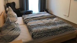 A neatly made bed with patterned bedding, a chair, a small table, and two wall-mounted paintings. - Beautiful barrier-free 3 1/2 room apartment near the beach!  (Allensbach)