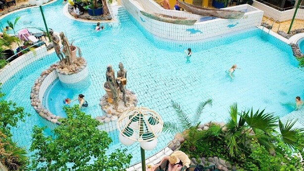 An indoor swimming pool with multiple sections, including a central area with a rock formation and a smaller enclosed area with a boat-shaped structure.
