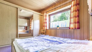 A bedroom with a wooden ceiling, a window with curtains, a bed with white bedding, and a wooden wardrobe. - 3-Bedroom Apartment  (Grömitz)