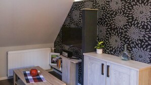 A compact kitchenette with a microwave, a small table with a checkered placemat and a red object, a cabinet with two doors, a potted plant, and a decorative wall with a floral pattern.