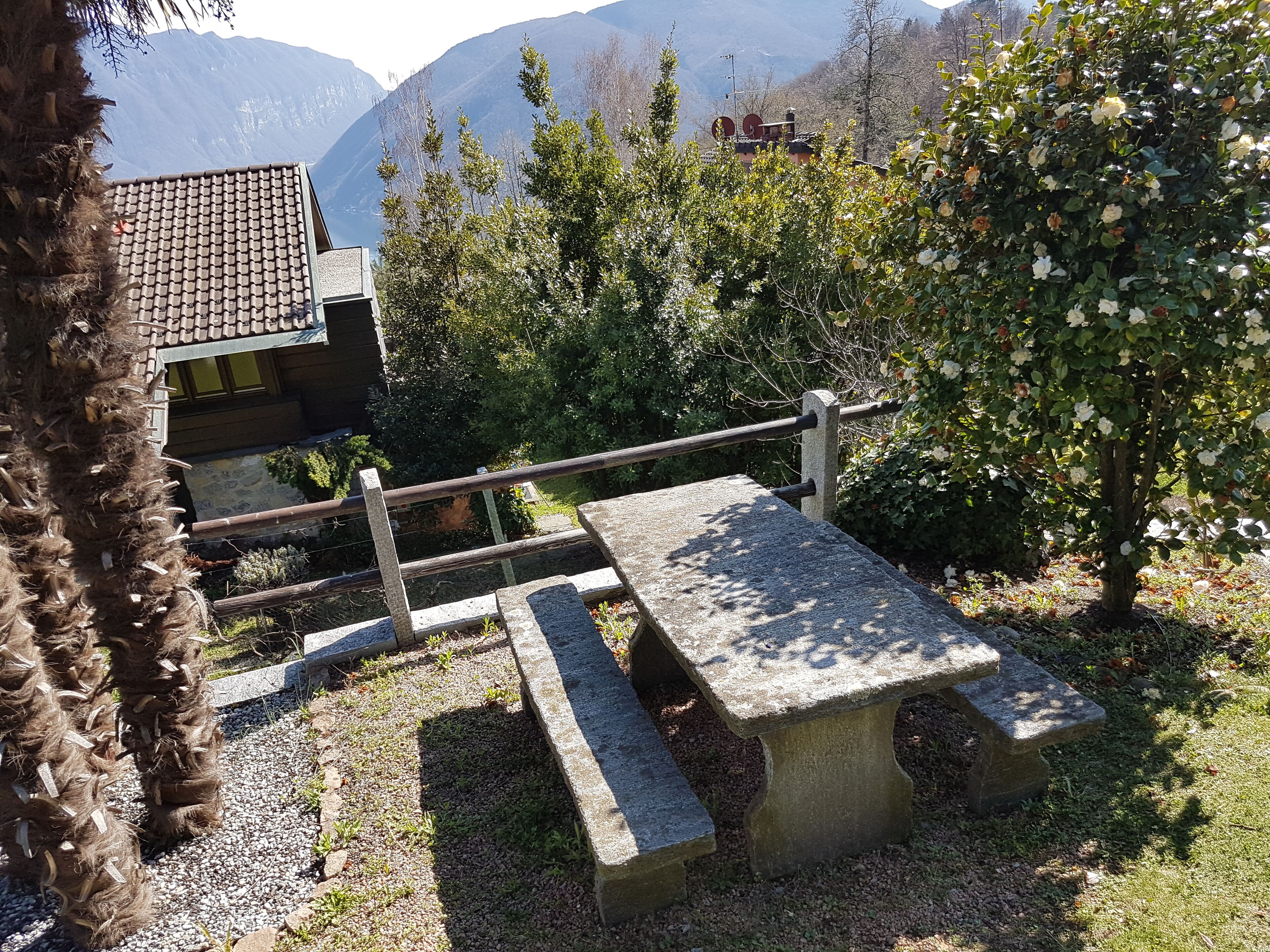 Eine Steinsitzbank mit Metallgeländer in einer Gartenanlage mit Bäumen und Blick auf die Berge im Hintergrund.