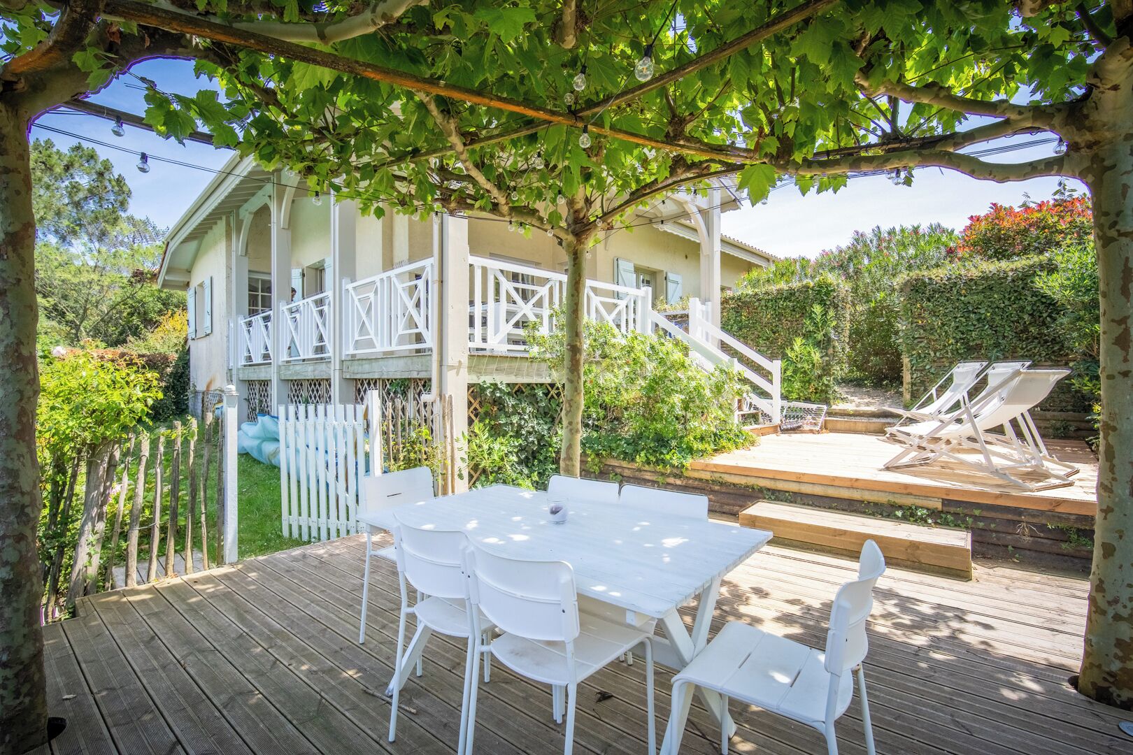 Un espace extérieur abrité, doté d’une terrasse en bois, de mobilier blanc et d’une pergola couverte de plantes grimpantes.