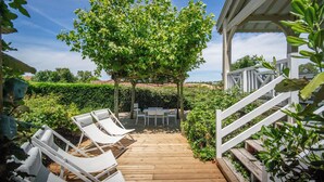 A wooden deck with white lounge chairs and a table, surrounded by greenery and a view of the sea.
