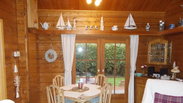 A wooden cabin interior with a dining table, chairs, a fireplace, and a view of the garden through large windows.