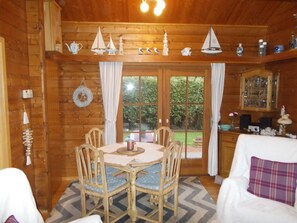 A wooden cabin interior with a dining table, chairs, a fireplace, and a view of the garden through large windows.