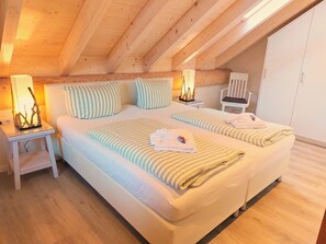 Un dormitorio con techo de madera, dos camas con ropa de cama a rayas, lámparas de noche, una silla y un armario.