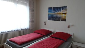 Tweepersoonskamer met twee bedden, elk met rode kussens en grijze beddengoed. Er is een radiator, een nachtkastje met een lamp en een aan de muur hangend schilderij van een zonsondergang boven de zee.